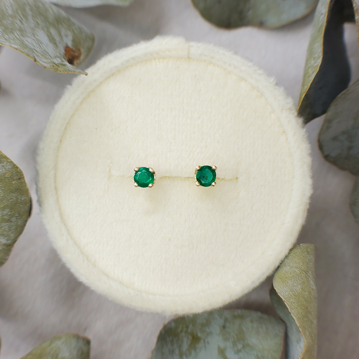 A vintage pair of small 14K yellow gold stud earrings with round green emerald gemstones, displayed on a round cream-colored jewelry box surrounded by dried green leaves.
